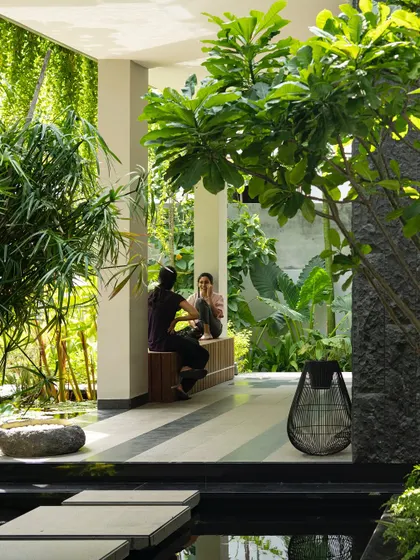 A quiet conversation nook on a covered veranda, looking out onto a water body and garden. The design uses built-in wooden seating and is surrounded by greenery, creating a space that feels both sheltered and open.