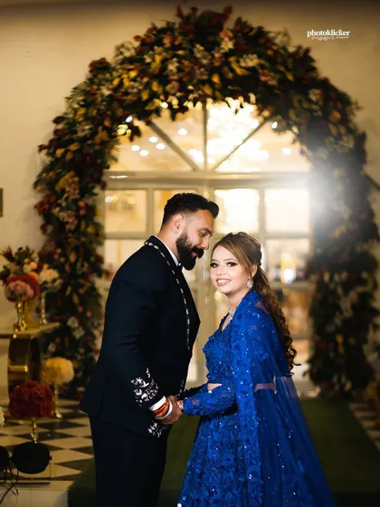 A couple holds hands under a beautiful floral arch, their matching blue and black outfits creating a coordinated and stylish look for their event.
