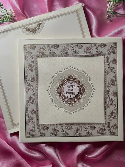 This elegant invitation suite features a soft, pastel color palette with a beautiful floral border. The central medallion with the couple's names adds a touch of classic charm, presented on a bed of pink satin to highlight its luxury.