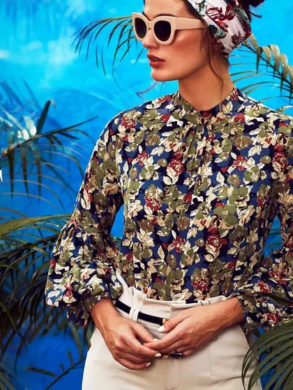 A tropical-themed editorial for 'SHECZZAR'. The styling features a vintage-inspired floral blouse, a headscarf, and statement sunglasses, creating a chic and colorful resort look.