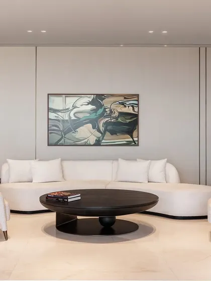 Minimalism and comfort converge in this serene seating area. A white curved sofa is paired with classic armchairs and a simple, bold coffee table. The abstract artwork adds a touch of color and movement to the otherwise tranquil space.