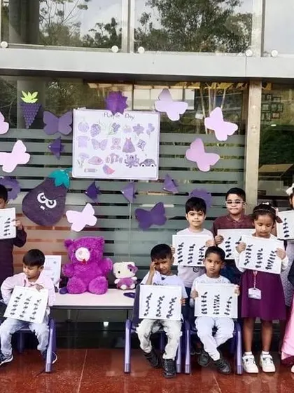 A class holds up their purple-themed artwork in front of a decorated wall, celebrating a day of colorful learning.