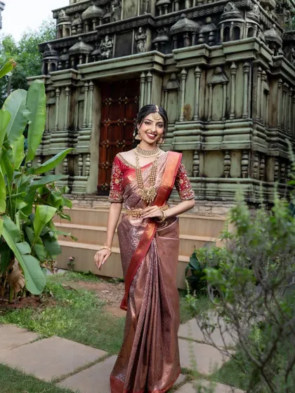A smiling bride-to-be in a bronze and red silk saree, set against a beautiful temple backdrop.