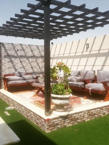 A traditional black pergola with a grid-style roof, providing a classic framework for an outdoor seating area. The base is finished with brick and stone, complementing the artificial grass surround.