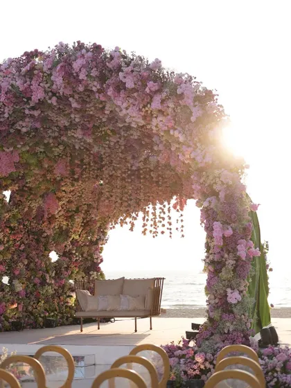 The sun sets behind a breathtaking floral mandap by the sea. The structure, covered in shades of pink and purple, is designed to catch the golden hour light, creating a truly magical and romantic setting for the vows.