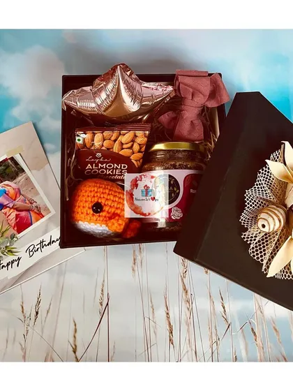 A stylish black gift box containing a personalized birthday card, almond cookies, a cute crocheted octopus, and other small treats.