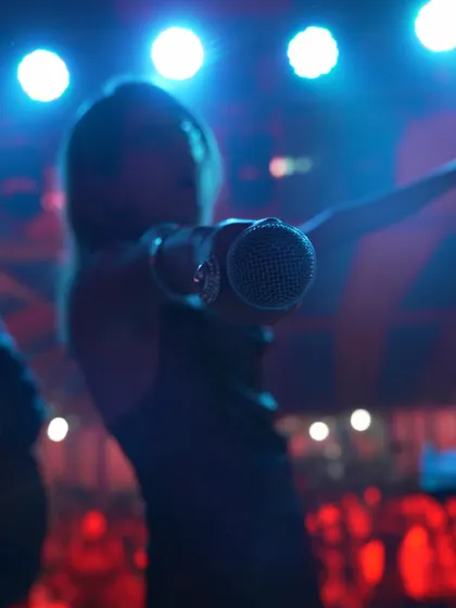 I'm holding the microphone towards the camera, with blue lights above me and the audience behind in this stage shot.