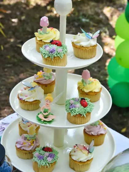 A tiered stand of beautifully decorated cupcakes for a fairy-themed party. Each cupcake is adorned with edible flowers, butterflies, ladybugs, or fairy figures.