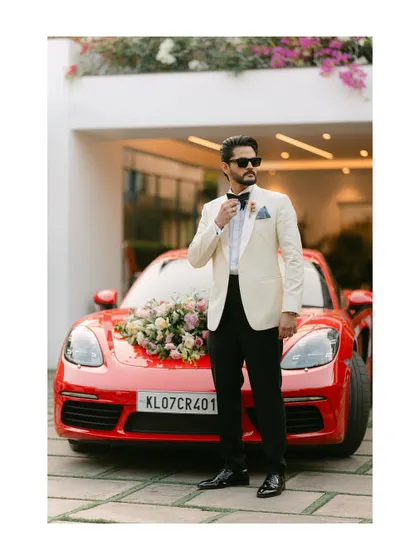 A stylish groom adjusts his bow tie, standing in front of a red Porsche decorated for the wedding. This image combines modern luxury with classic wedding elegance.