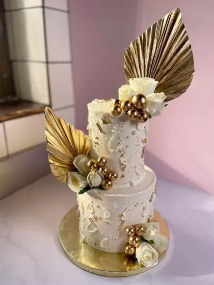 A modern and chic two-tiered custom cake. This design features a clean white buttercream finish accented with gold palm leaves, white roses, and delicate gold spheres for a touch of glamour.