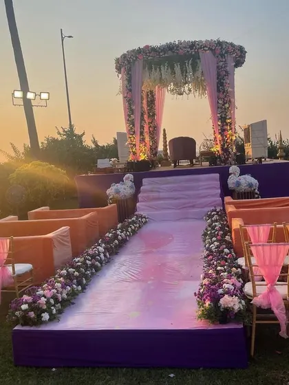A beautiful sunset view of a wedding mandap set up on a lawn overlooking the sea. The purple platform and orange seating add a pop of color to the romantic evening ambiance.