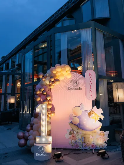 A unique photo corner featuring a large marquee '1', a custom-illustrated fairy house prop, and a personalized sign for the birthday girl, Raaina.