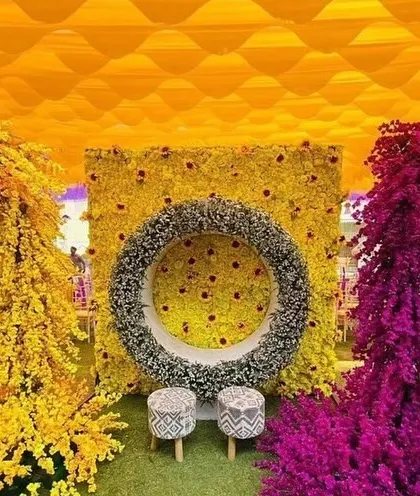 A creative and colorful photo point for a Haldi ceremony. This setup features a large floral wreath against a bright yellow backdrop with cascading yellow and purple flowers, perfect for guest pictures.