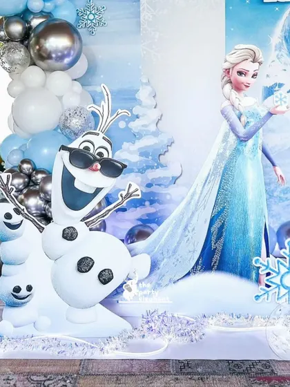 A close-up of the Frozen party decor, showing a life-sized Elsa cutout and a very cool Olaf wearing sunglasses, surrounded by snowmen props and a blue and silver balloon arrangement.