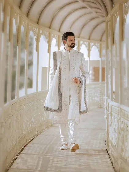 A portrait of the groom in his elegant white sherwani, walking through a sunlit palace corridor, capturing a moment of quiet confidence before the ceremony.
