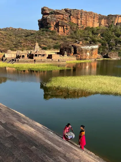 Two women in colorful sarees wash clothes by the river, with ancient temples and rocky hills in the background. This image beautifully blends the daily life of today with the history of the landscape.