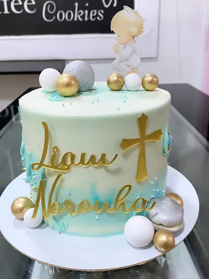 A serene and beautiful cake for a baptism celebration. The light teal and white cake is decorated with a gold cross, the child's name in a gold script, and a praying angel topper.