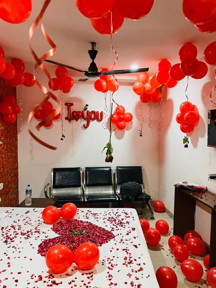 A hotel room transformed for a romantic surprise. The decor includes red balloons on the ceiling, rose petals on the bed, and 'I love you' foil balloons on the wall.