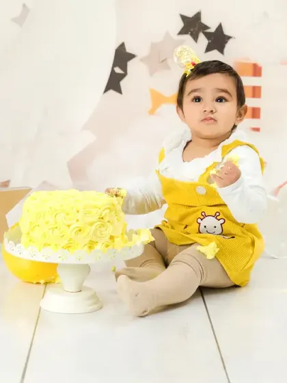 A baby girl in a yellow outfit sits next to her birthday cake in a dreamy, star-filled setup. A sweet and gentle cake smash photo.