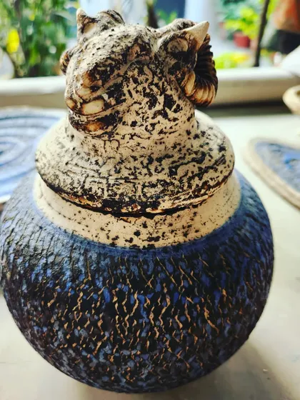 This decorative jar was finished using an Obvara firing technique, which creates the distinctive crackled and colored surface. The lid is sculpted to resemble a ram's head, adding a powerful artistic detail.