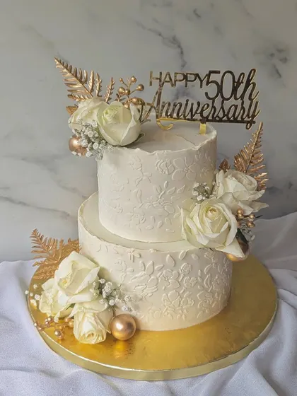 Celebrating a golden anniversary with this elegant two-tier cake. The design features a subtle floral stencil, white roses, and striking gold-dusted ferns for a touch of glamour.