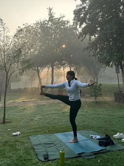 Holding a standing leg raise against a backdrop of sunlit trees.