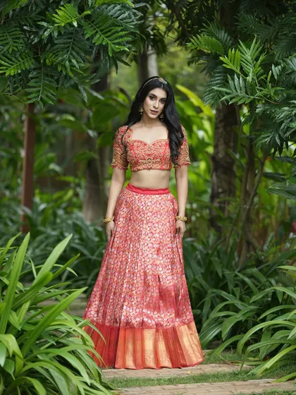 This full-length shot captures the grandeur of the coral-red half saree, an ideal choice for a bride's main ceremony or reception.