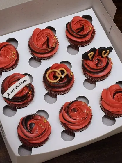 Celebrating new beginnings with this custom engagement cupcake box. The set features rich red velvet cupcakes with red and black frosting, accented with edible gold dust, and detailed fondant work like intertwined rings and custom initials.