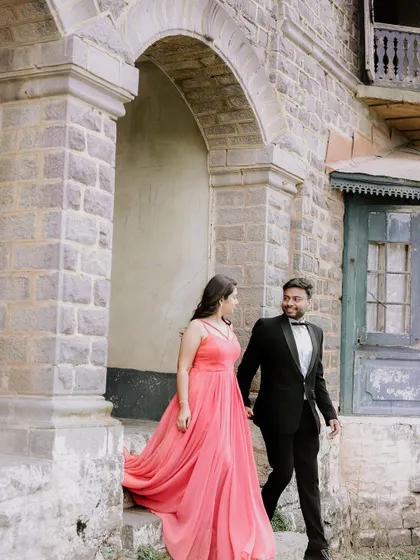 A couple dressed in formal wear emerges from a rustic stone archway. The contrast between the elegant attire and the old-world charm of the location creates a timeless pre-wedding photograph.