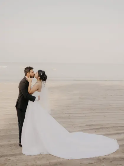 The couple shares a kiss against the vast expanse of the beach and sky, a beautiful destination wedding portrait.
