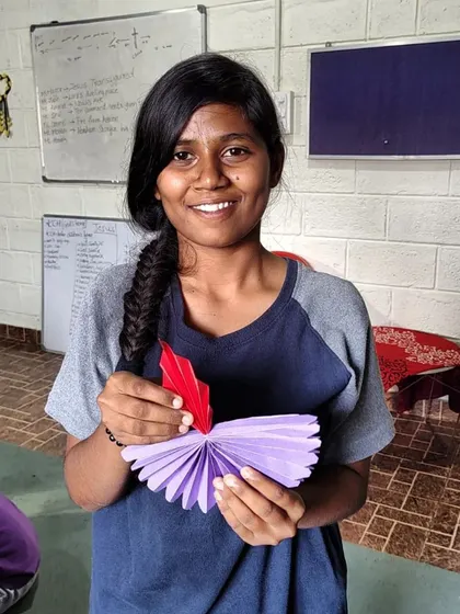 A smiling girl holds up her purple and red paper diya. Each creation is a small beacon of the light and hope we celebrate during Diwali.