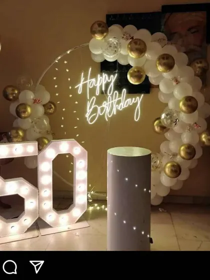 An elegant and minimalist 50th birthday setup. It features a semi-circle of white and gold balloons on a ring frame, a neon "Happy Birthday" sign, and large marquee "50" numbers.