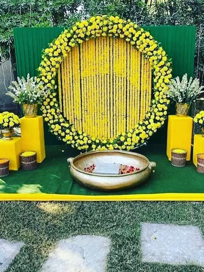 An outdoor Haldi ceremony setup with a circular backdrop of hanging yellow flowers. The surrounding green lawn and simple props create a fresh and natural setting.