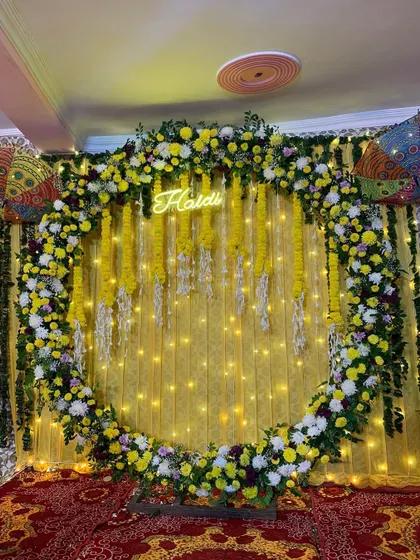 A close-up of a stunning circular floral arch for a Haldi ceremony. The arch is densely packed with yellow and white flowers, framing a custom 'Haldi' neon sign for a modern and festive photo opportunity.