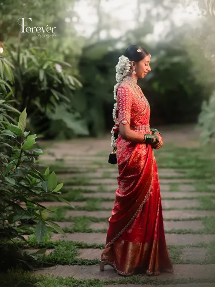 A full-length shot of the bride in a garden setting, her red saree standing out beautifully. The look is traditional, elegant, and timeless.