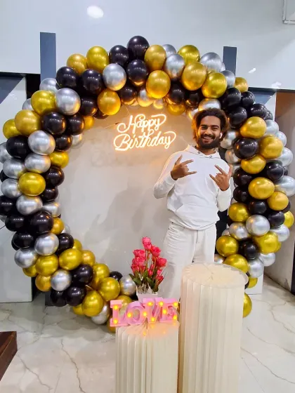 A customer posing with the black, gold, and silver balloon ring. This shows the scale of the backdrop, which features a 'Happy Birthday' neon sign.