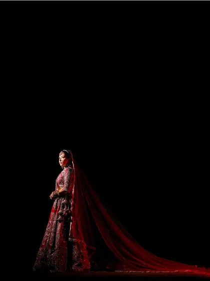 A breathtaking full-length shot against a black background. The bride's red lehenga with its long trail creates a silhouette of pure drama and elegance.