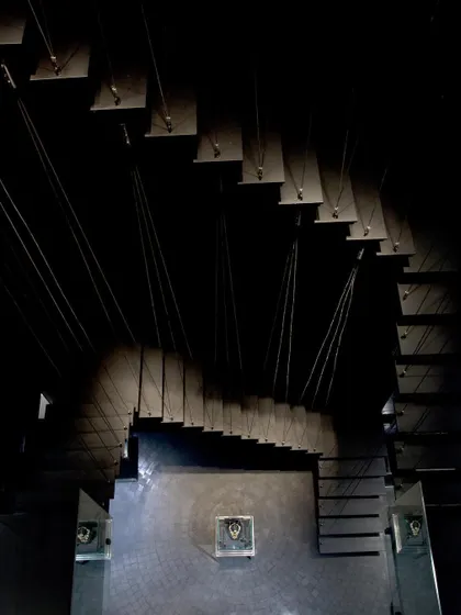 A view looking up the suspended staircase at the ORRA store. The design, with its thin metal treads and cable supports, creates a sense of lightness and drama, drawing the eye upwards through the dark, cavernous space.