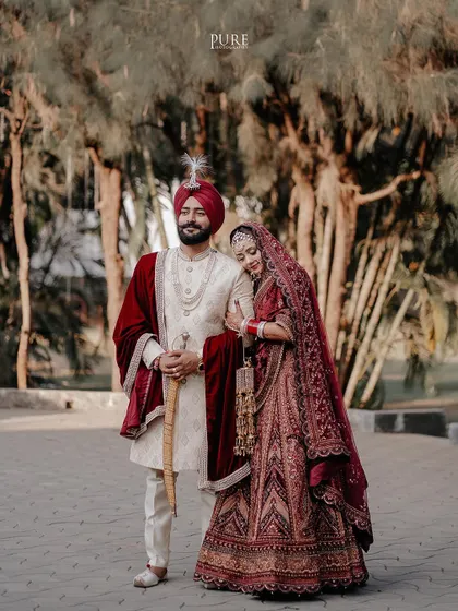 A royal portrait of the bride and groom. The rich maroon velvet and intricate embroidery of their outfits are captured in stunning detail.