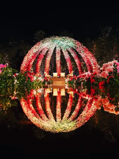 A stunning nighttime view of the open-air floral dome mandap. The structure is lit to create a glowing sphere of red and white, reflecting beautifully in the water for a truly magical wedding moment.