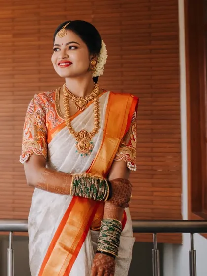 A full-length shot of the bride in her off-white and orange saree, looking happy and ready for her wedding.