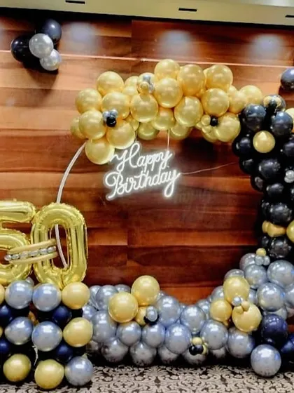 A 50th birthday celebration featuring a half-arch balloon arrangement in gold, black, and silver, with a neon sign and large foil number balloons.
