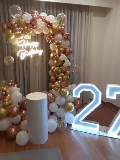 A 27th birthday party with a rose gold, gold, and white balloon arch. The setup includes a "Happy Birthday" neon sign and large marquee numbers.