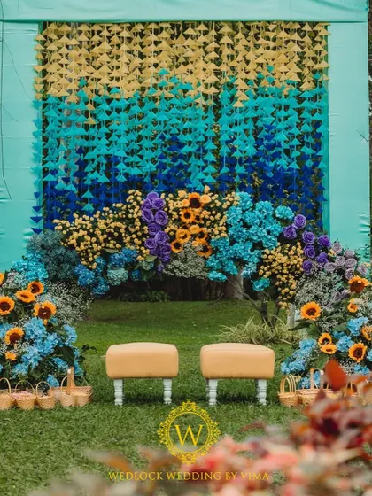 A vibrant floral backdrop and welcome sign for a colorful Haldi and Mehendi ceremony. The mix of orange, blue, purple, and yellow flowers created a stunning photo opportunity for the couple and their guests.