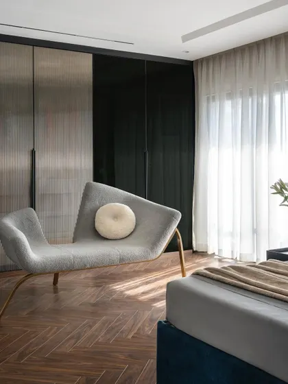 A detail from the master bedroom in the Evelyn penthouse. The custom grey chaise lounge with its unique asymmetrical design sits in front of sleek, metallic-finish wardrobe shutters, showcasing a blend of comfort and high-end design.