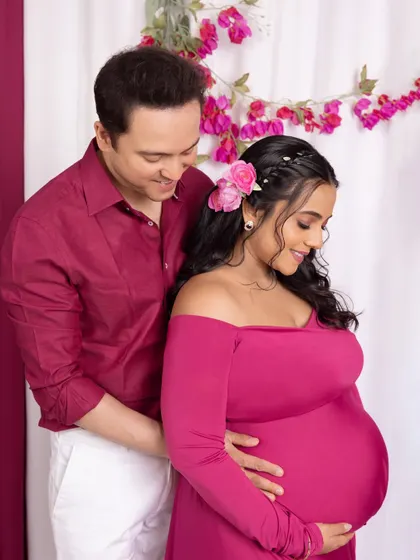 A candid moment during a floral-themed maternity shoot. The father-to-be smiles down at his partner as she cradles her belly, surrounded by vibrant pink flowers that add a touch of romance.