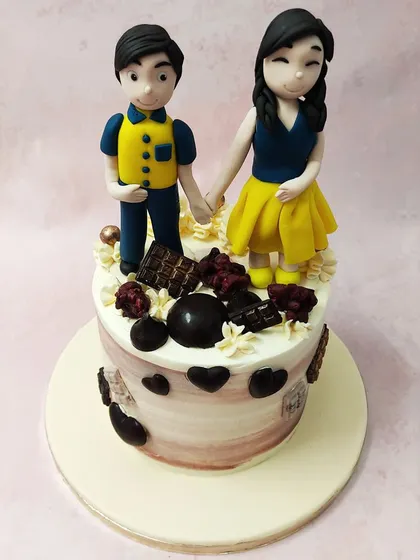 We celebrate partnership with this sweet couple anniversary cake. A custom-made topper of the couple holding hands sits atop a cake decorated with chocolate hearts and bars, creating a personalized and romantic tribute to their love story.