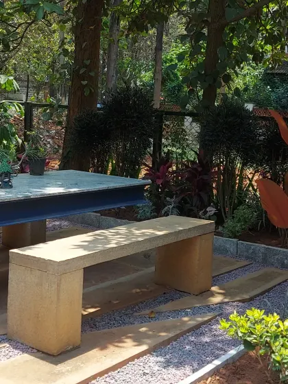 A secluded outdoor dining area with a concrete table and stone bench. The space is nestled among trees and features a gravel ground cover, creating a rustic and natural feel.