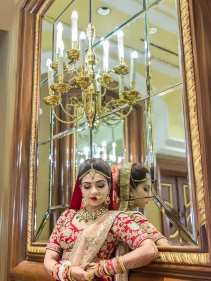 A reflective bridal portrait. The bride, dressed in a stunning red lehenga, poses against a large, ornate mirror, creating a sophisticated and artistic image.
