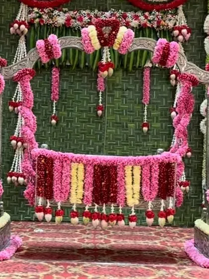 A detailed view of one of my silver cradles (thottilu) available for rent, decorated for a naming ceremony. I've adorned it with dense garlands of pink and red roses against a backdrop of woven banana leaves, perfect for a traditional event.